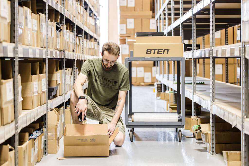 Warehouse worker with mobile barcode scanner kneeling down in a warehouse aisle and looking into a box, demonstrating picking and packing best practices for the warehouse