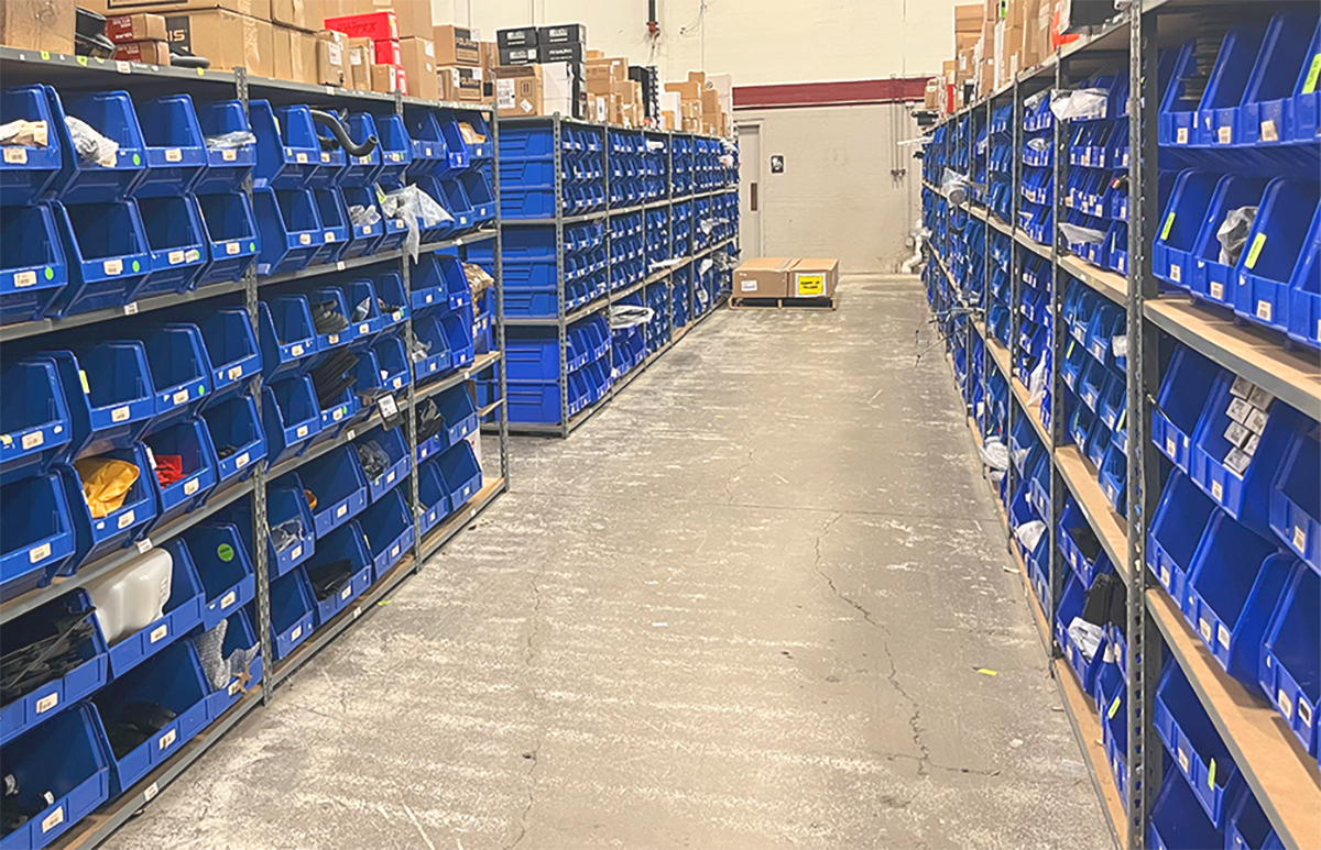 A warehouse aisle with shelves and blue bins full of products arranged in a pick face. There is a break in the pick face for workers to cut through to the next aisle.