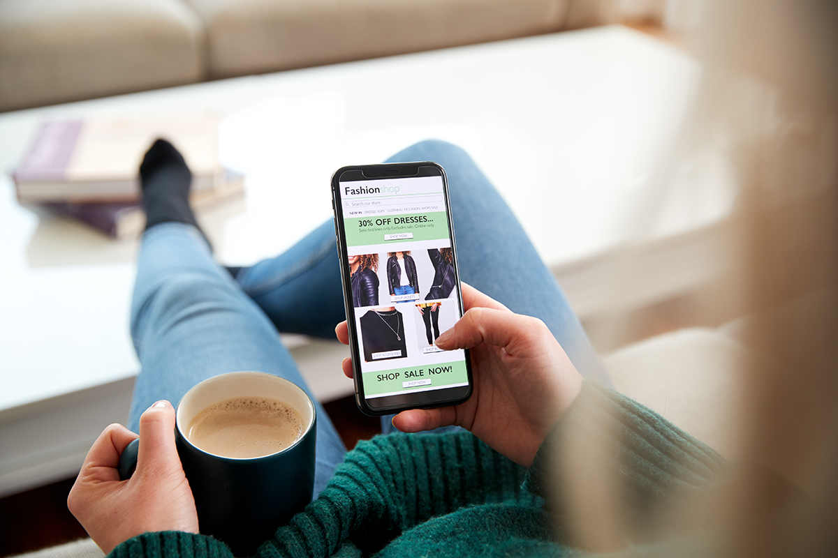 An ecommerce shopper is holding a mobile device and a cup of coffee, scrolling a fashion website with women’s apparel.