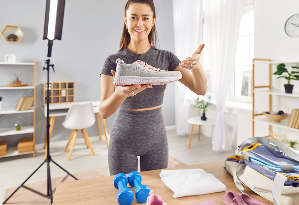 A female fitness influencer wearing athletic clothes stands in a bright, well-lit office holding an athletic shoe while filming a TikTok video for social media selling.
